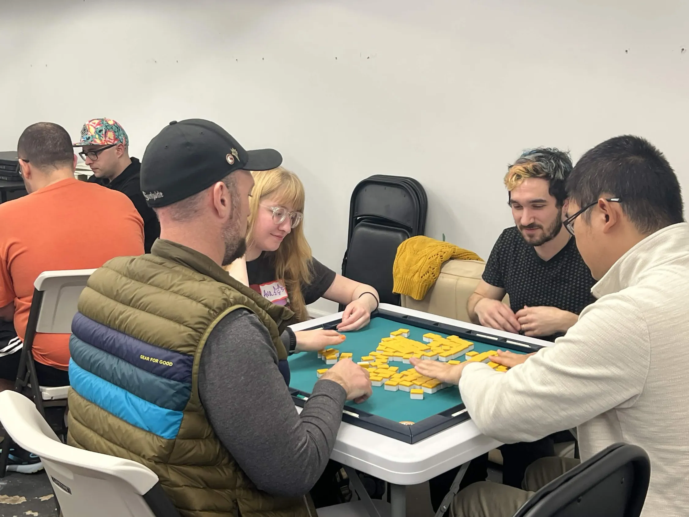 Four players sort yellow mahjong tiles at the start of a hand. One player smiles and wears a name tag while tiles are being set up on the green mat.