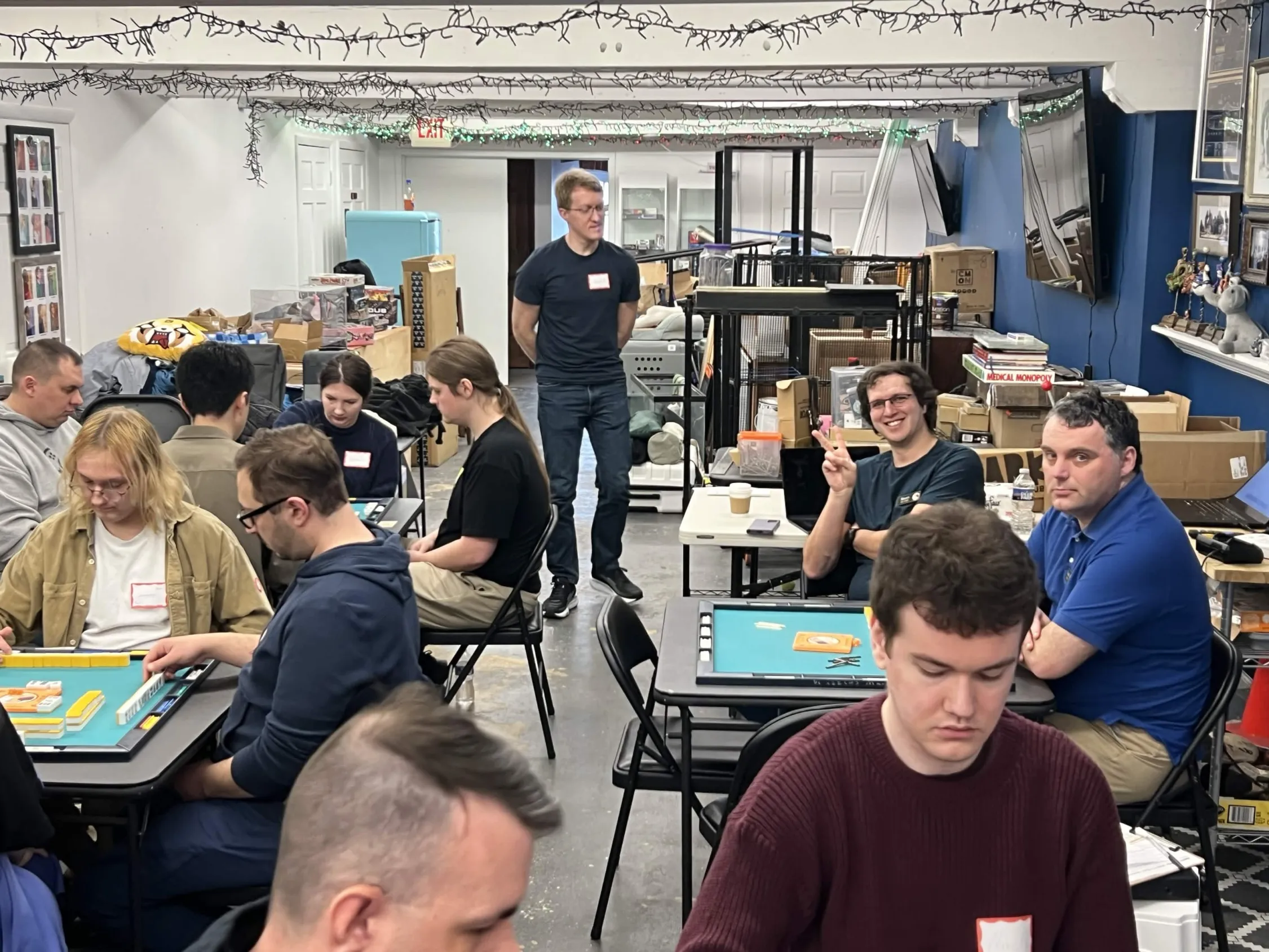 A tournament organizer stands in the center of the room overseeing active play at Omnihedral. Players study their tiles in the foreground while more tables are visible in the background.