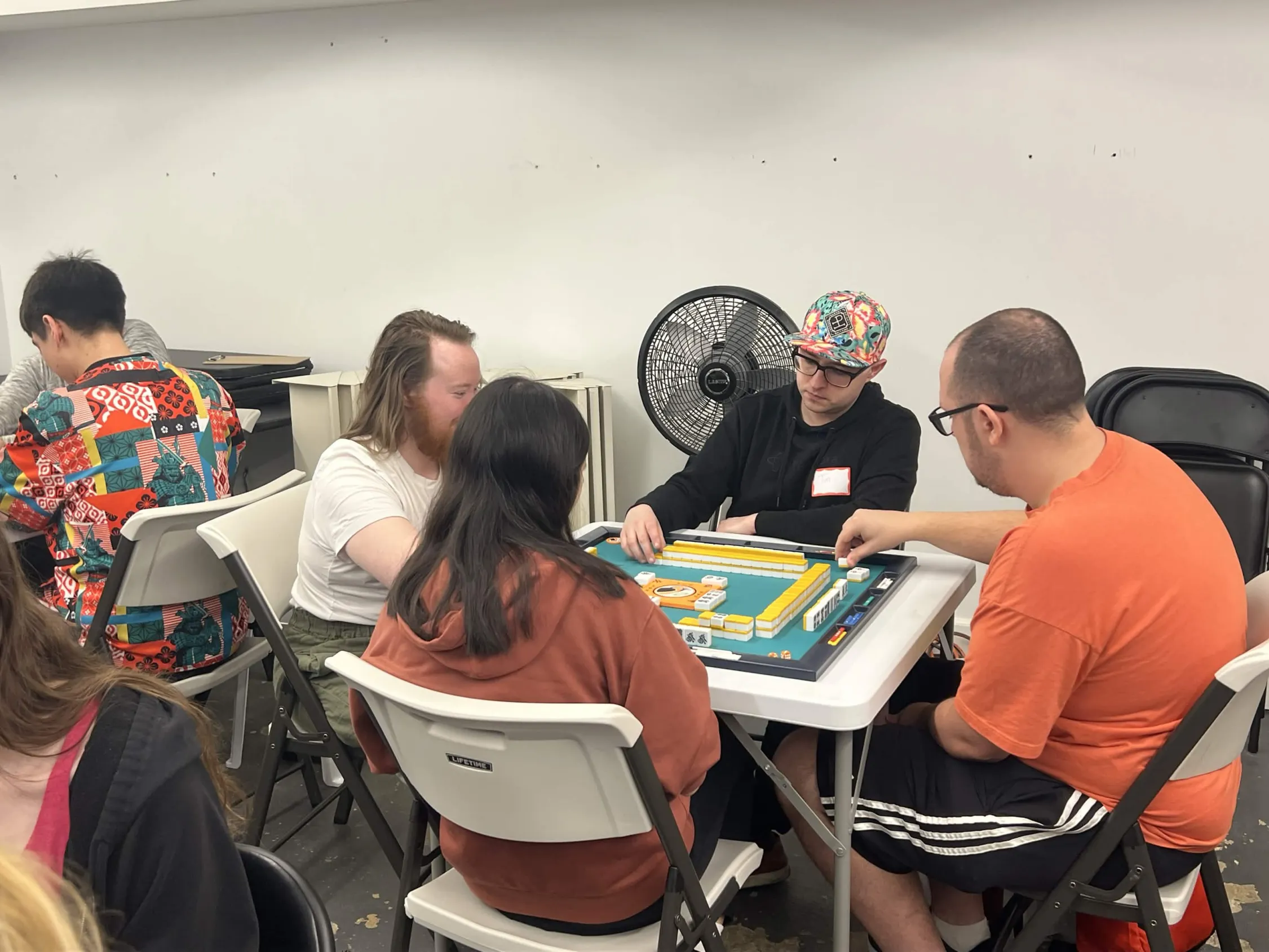 Four players concentrate on an active mahjong hand. A player wearing a colorful floral cap leans in to study the yellow tiles arranged on the table.