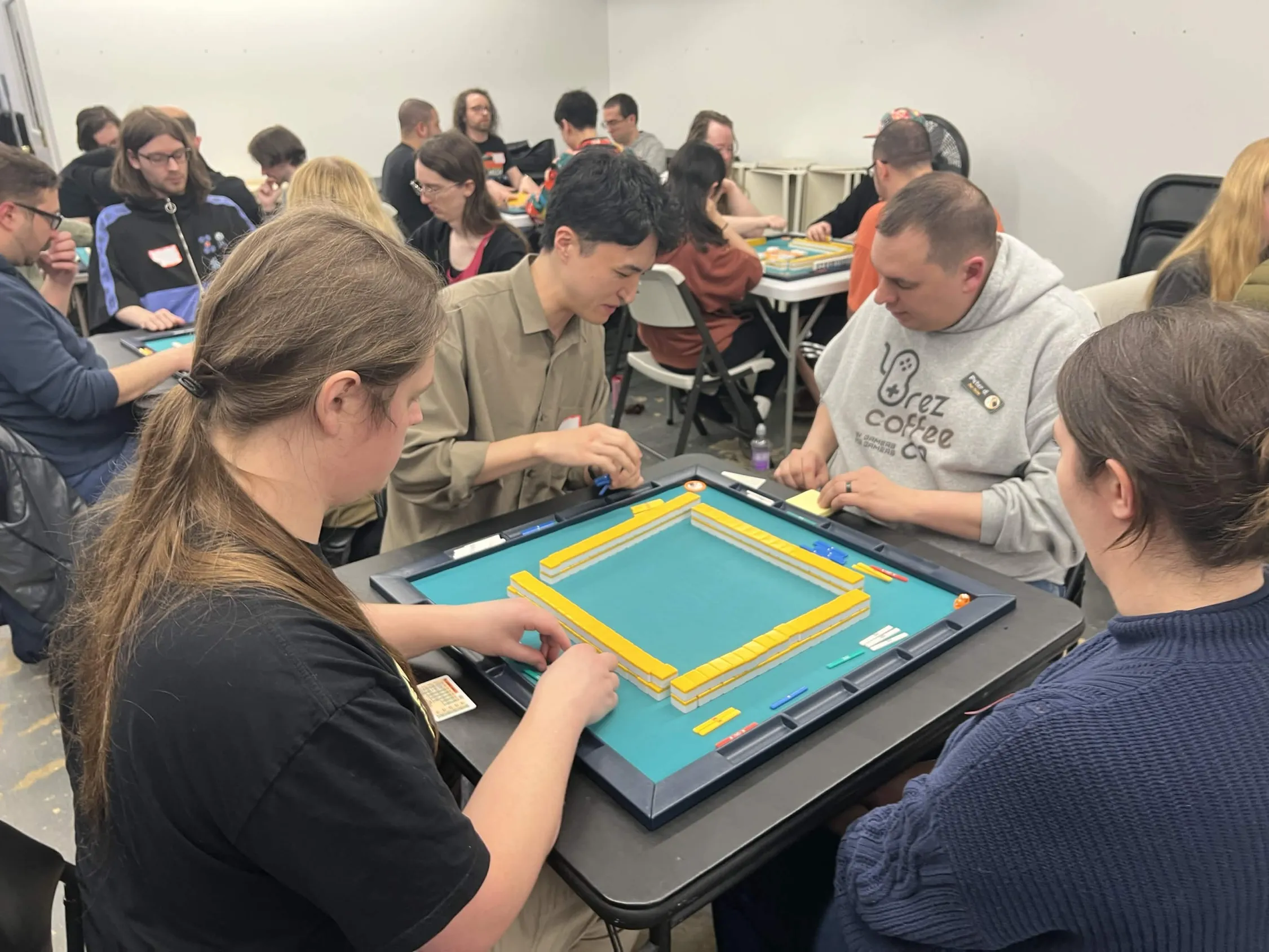 Players build their starting walls at a large mahjong table while a full room of competitors does the same in the background. The busy March 2026 tournament floor at Omnihedral stretches across multiple tables.