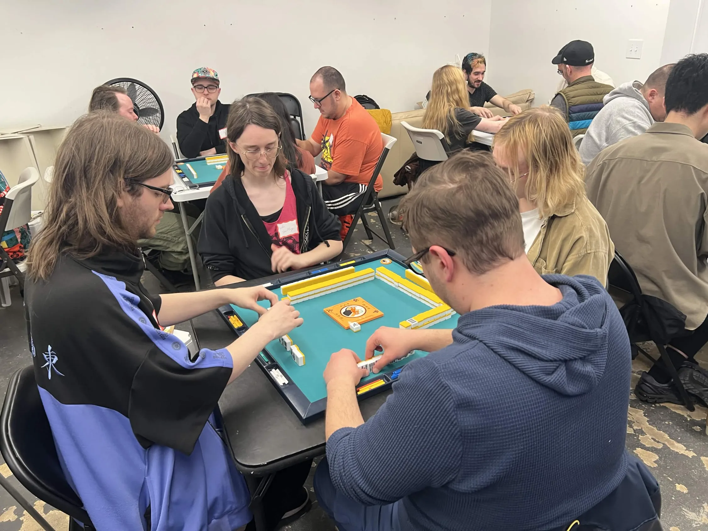 Four players lean in over a green-matted mahjong table as one makes a discard. Multiple tournament tables and players are visible in the background at Omnihedral.
