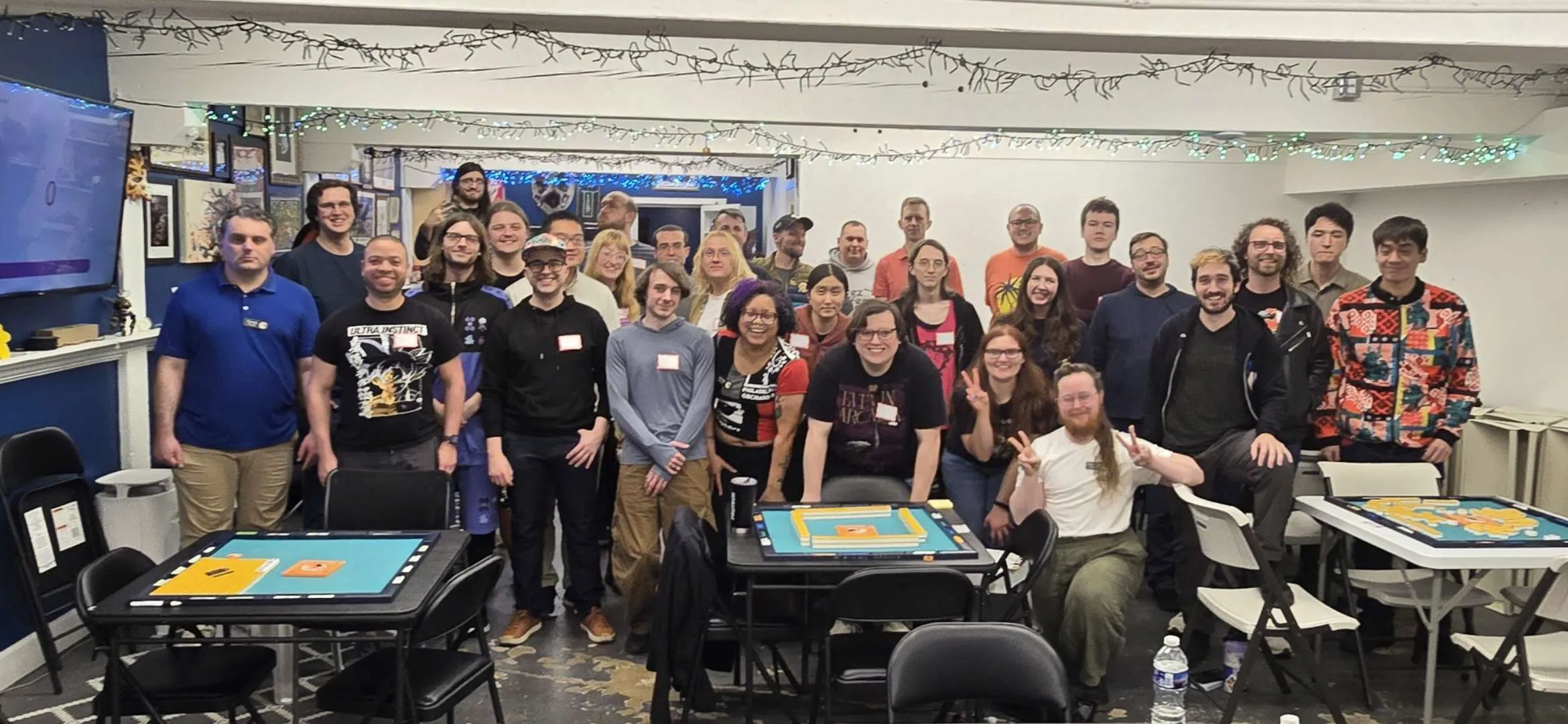 Group photo of approximately 30 tournament participants gathered at the end of the March 2026 one-day event at Omnihedral game store in Savage, MD. Mahjong tables are visible in the foreground.