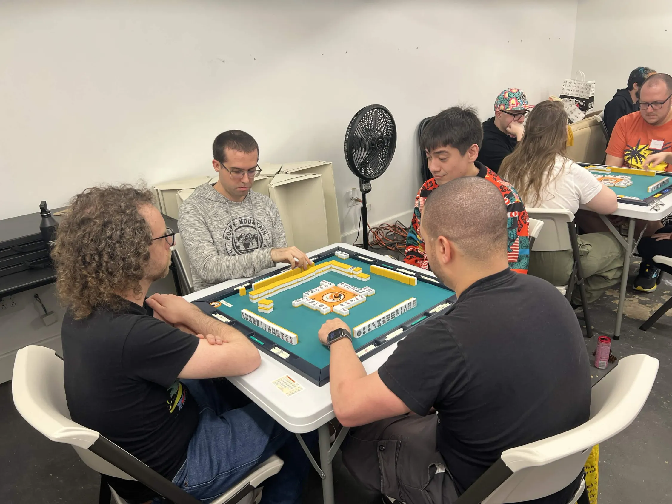 Four players sit in focused concentration around a mahjong table mid-game. Tiles are arranged on the mat as players study their hands carefully.