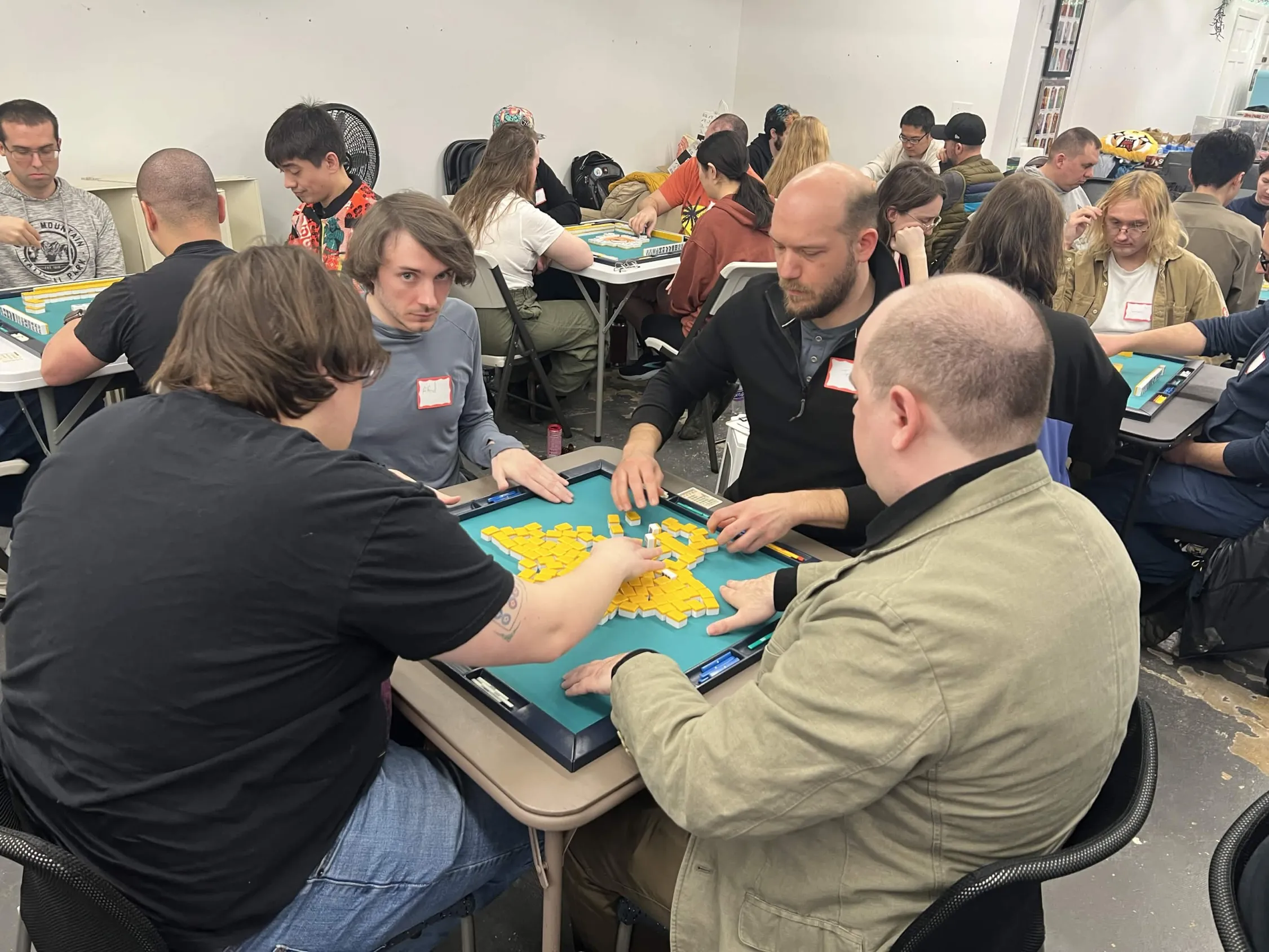 Players shuffle and mix yellow mahjong tiles across the mat at the start of a new hand, with a busy room of other competitors visible in the background.