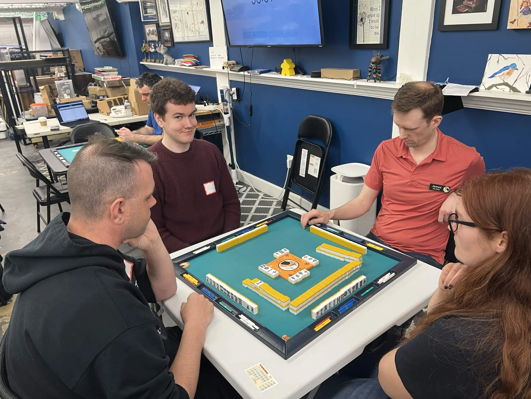 A young player in a maroon sweater smiles during a tournament game at Omnihedral. Three other players are seated around the table, with the store's blue wall and shelves visible in the background.
