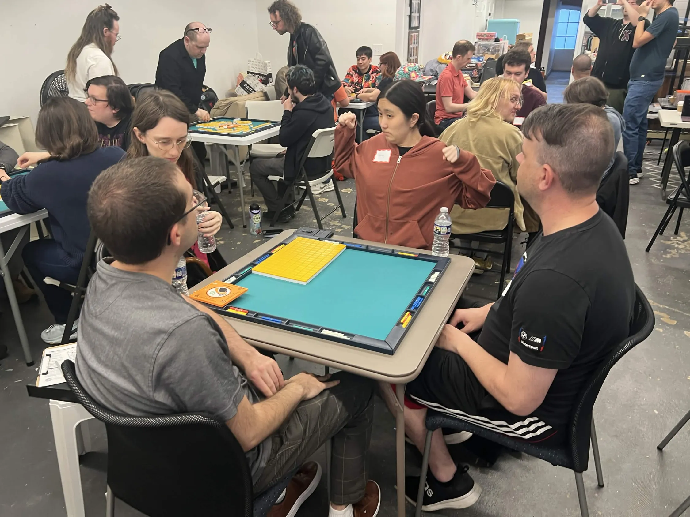 A player reaches across the mahjong table to make a tile claim while three opponents watch. Several other games are underway in the background at Omnihedral.