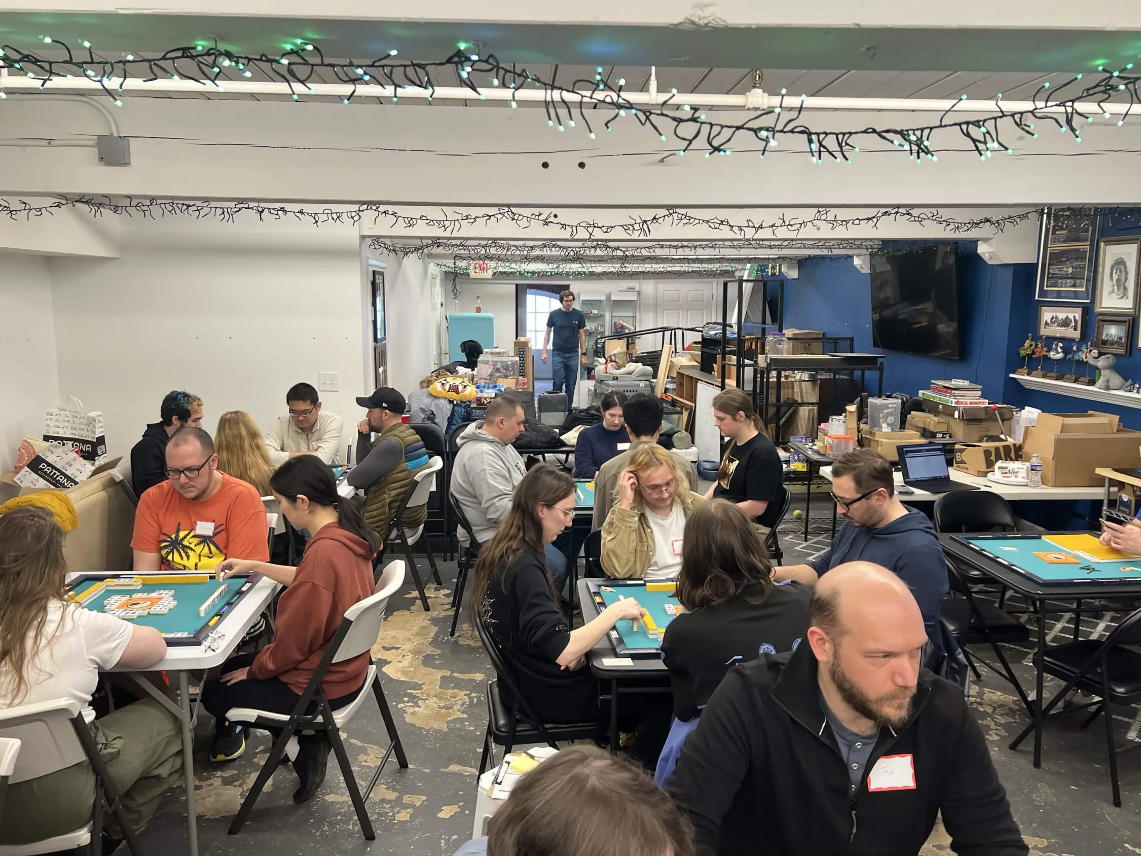 Wide view of Omnihedral's game room packed with players at mahjong tables during the March 2026 one-day tournament. String lights hang from the ceiling across the full venue.