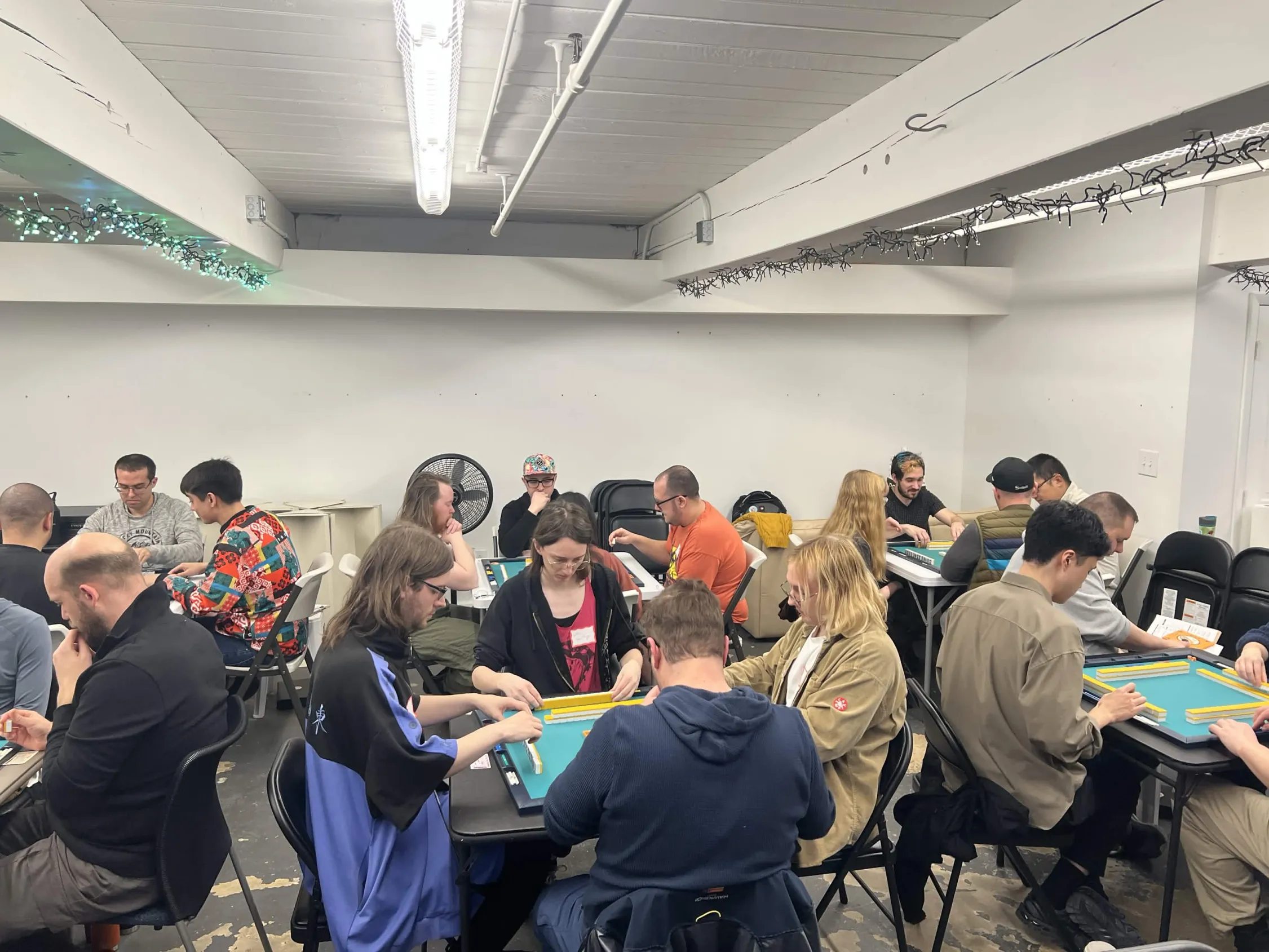 Wide shot of the full tournament room showing players at multiple mahjong tables in active play. The room is packed for the one-day event at Omnihedral.